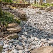 Dry streambed lined with smooth stones and surrounded by greenery.