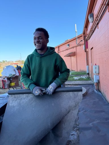 A smiling person in a green hoodie working with construction equipment outdoors.